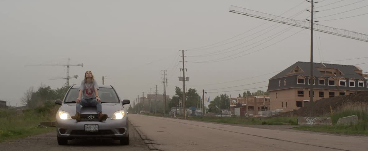A woman sits down on her car hood as its parked along the shoulder of a rural two lane road
