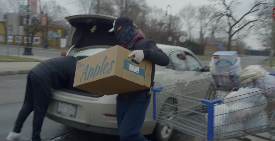 A man wearing gloves and a bandanna over his face carries a cardboard box from a shopping cart to the trunk of a car where another is making room