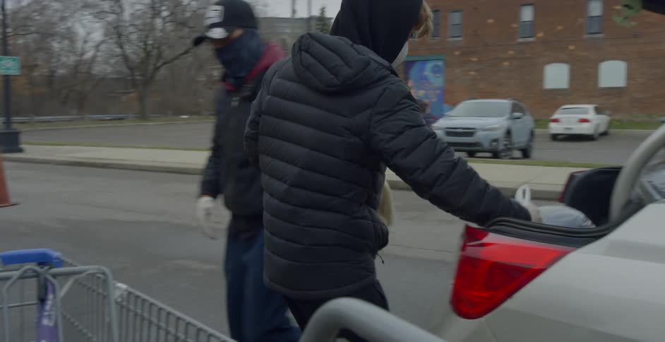 Two men wearing face masks load groceries into the back of a car trunk