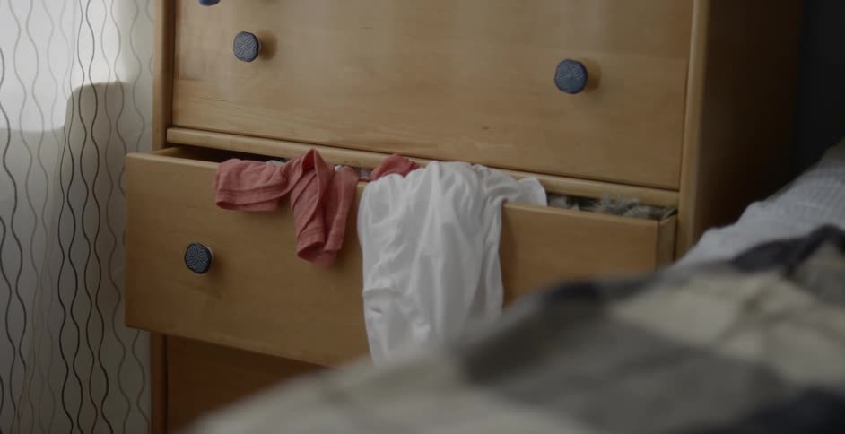 A dresser drawer cracked open with clothing spilling out next to a bed