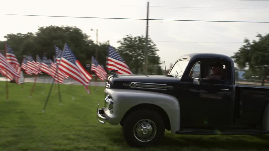 An old pickup truck drives onto a lawn filled with american flags