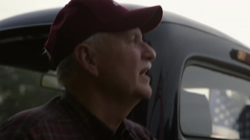 An older man holds a flag as he stands near his truck