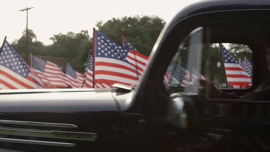 An older man sits in a well maintained old pickup in front of a lawn covered with american flags