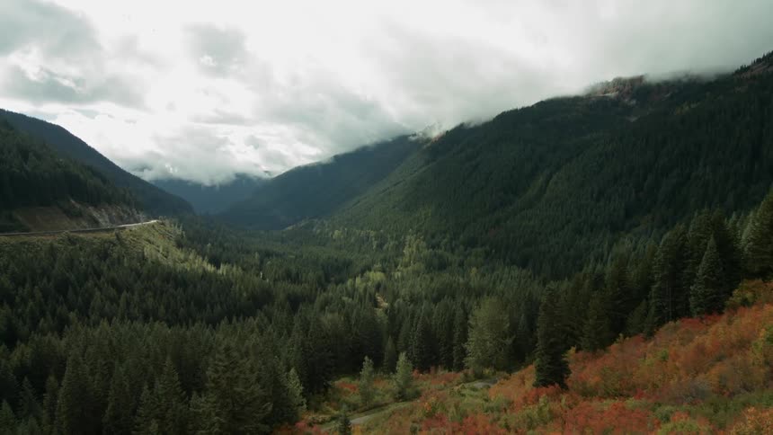 Aerial footage of a winding mountain road, forest, snow-capped peaks and low-lying clouds