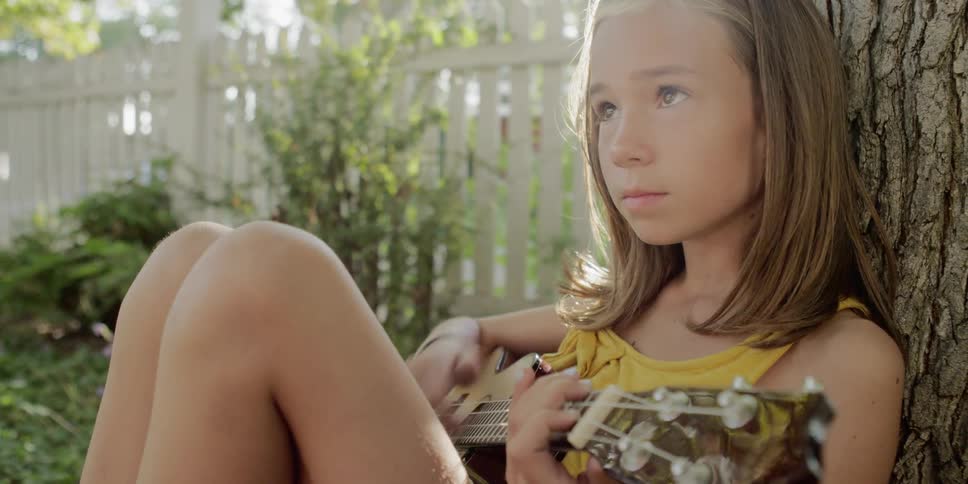 A young girl sits against a tree and strums a guitar