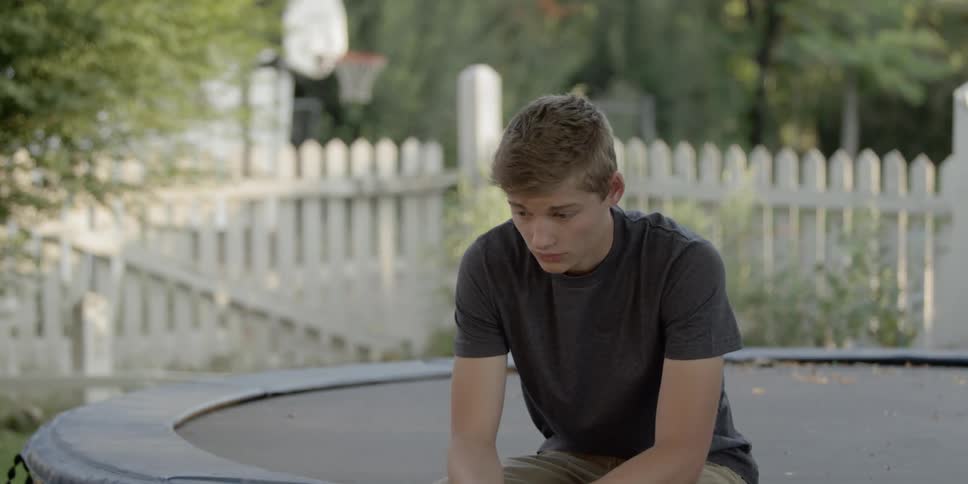 A teenage boy sits on the edge of a trampoline in the backyard looking sad