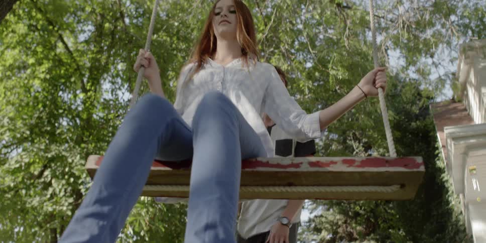 A young man pushing a young woman on a wooden swing in the yard of a house