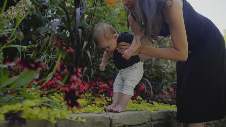 A young woman in a dress leans over as she playfully helps her baby boy stand on the ledge of a planter