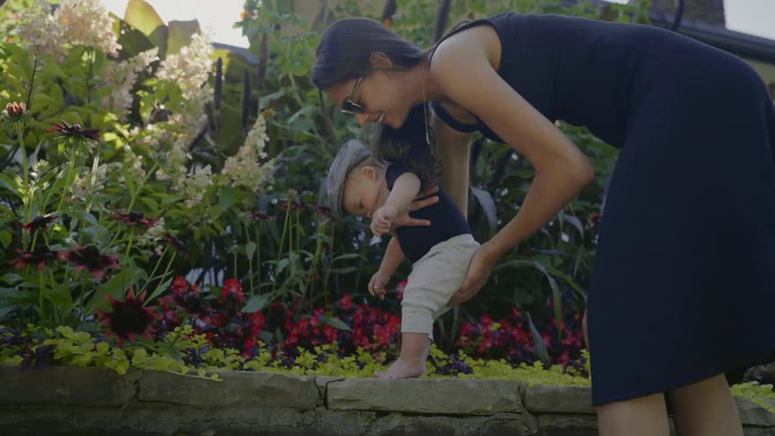 Mother holding her baby upright so their feet are touching a stone wall of a flower bed as they try to teach the baby how to walk