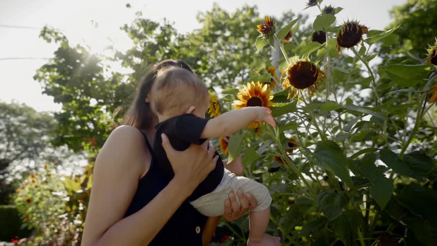 Woman holds baby touching sunflower in the public park