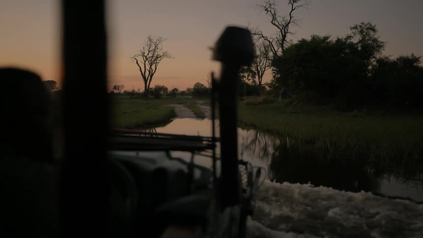 A jeep splashes as it travels through water with grass on either side at sunset