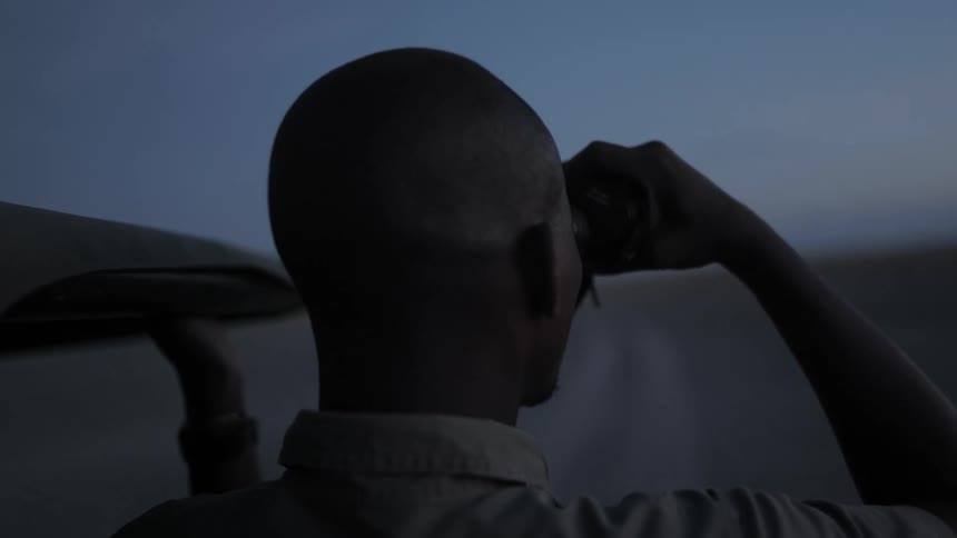 Male from the military is standing next to a vehicle while looking through binoculars along a dirt road
