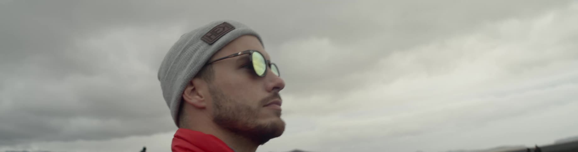 Man wearing a toque and sunglasses looks around as he stands under the cloudy sky