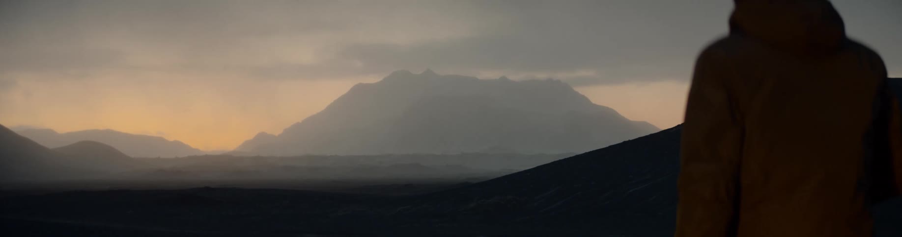 A person in a winter coat and ski mask walk out on a ridge with mountains and clouds on the horizon