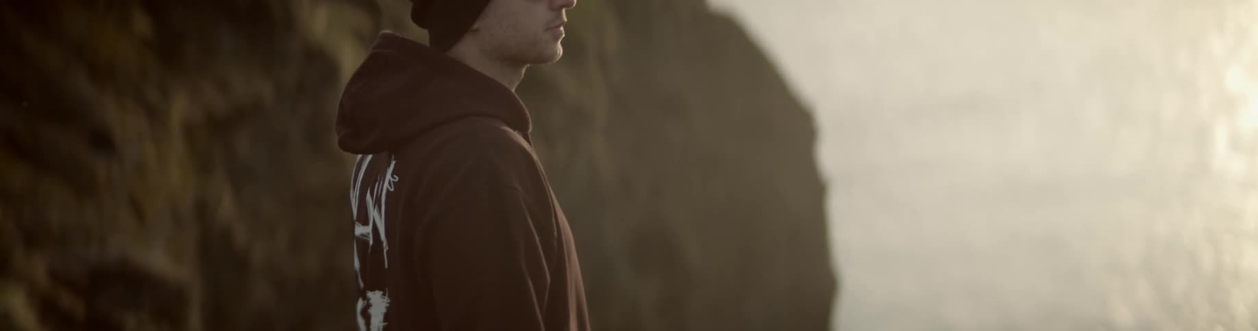 Man wearing a beanie and sunglasses stands on the edge of the cliff at sunset