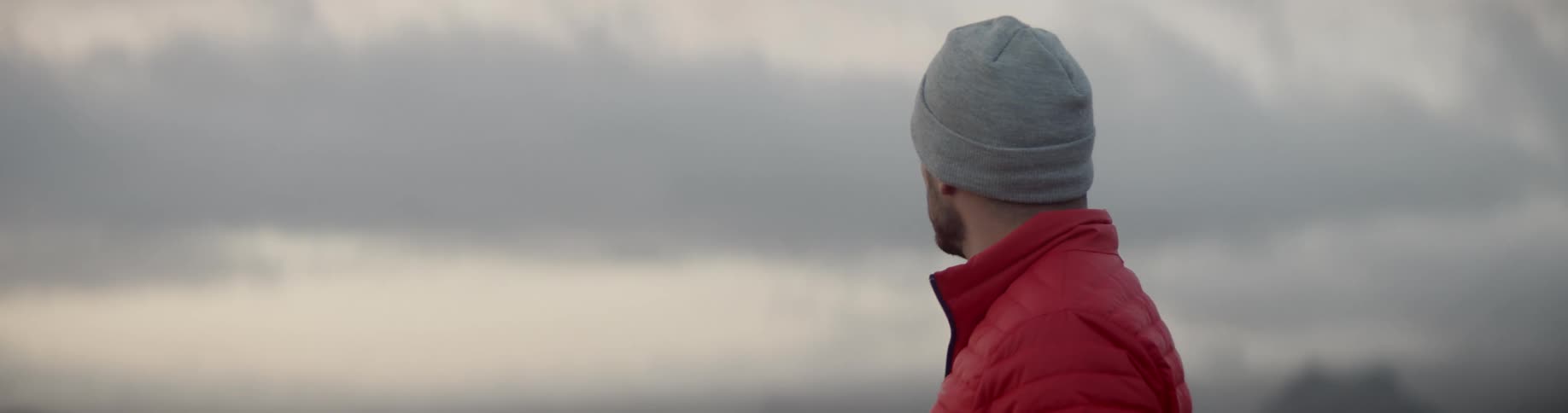 A man in a gray beanie hat and red quilted jacket looks out at the cloudy sky