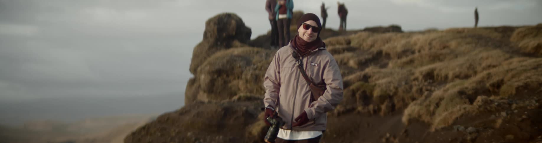 A man takes a photograph of the view while standing on a cliff then turns and climbs back up the hill