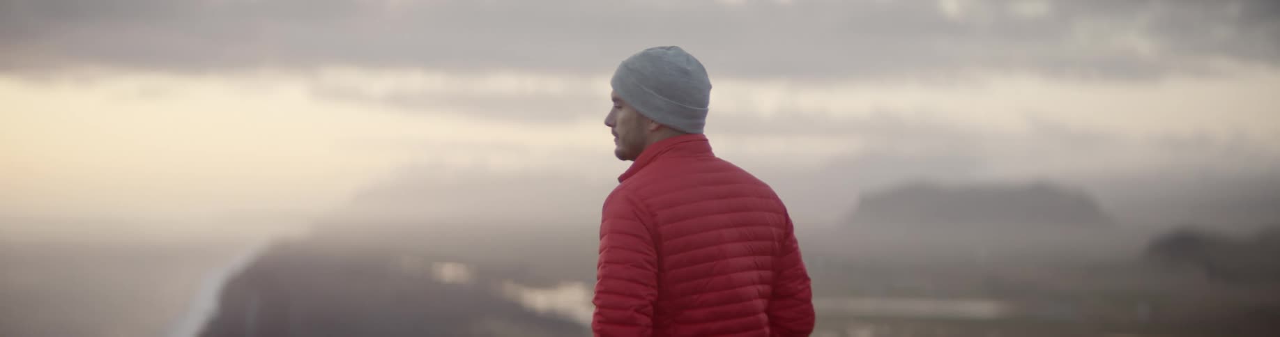 Man wearing a red coat and grey beanie stands on the edge of a cliff