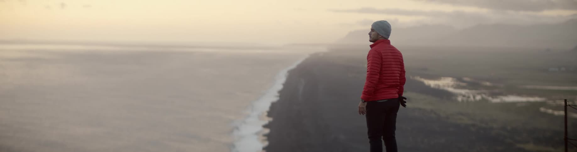 A man in cold weather apparel stands on a mountain high above a long shoreline