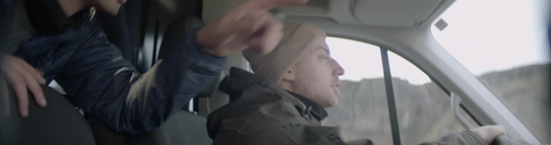 A man rides in the back of a van and leans over the seat to talk to the driver