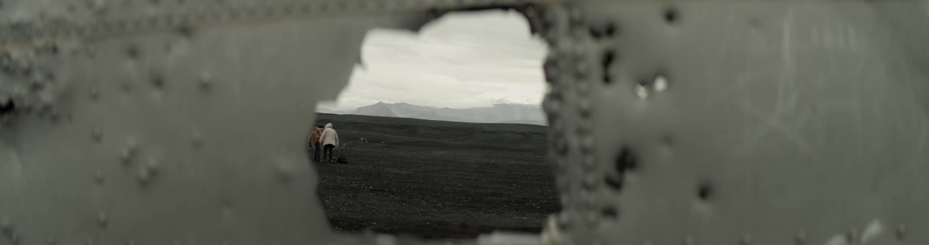 People are viewed on a barren landscape through a jagged hole in the metal of a wreck