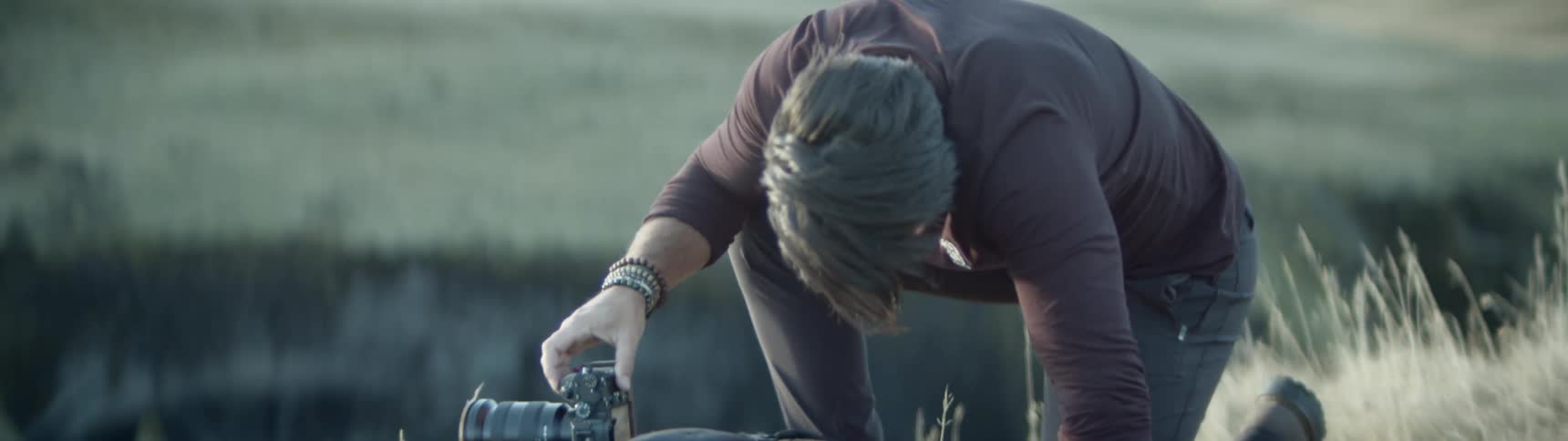 Man leans over dlsr camera to adjust settings in grassy field