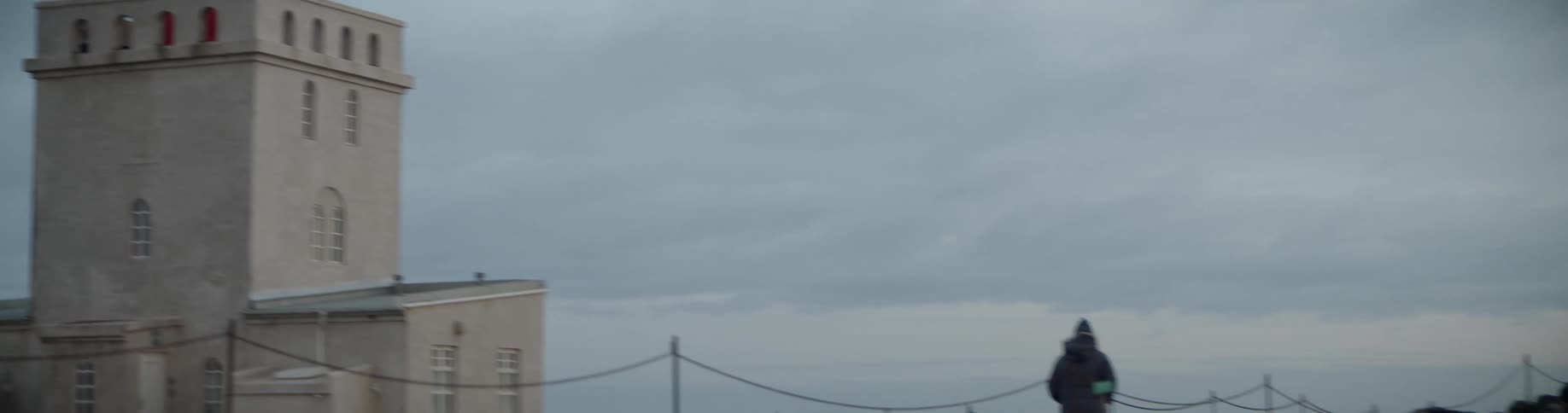 A person walks along a path next to a small lighthouse tower located near the ocean