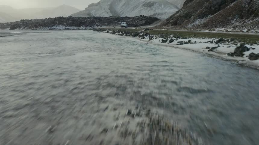 An aerial view of a truck traveling on a road close to a rocky shore with mountains on a cloudy day