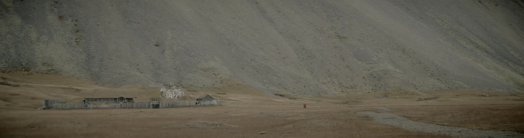 Wooden buildings surrounded by a wooden fence on a plain at the base of a gravelly mountain