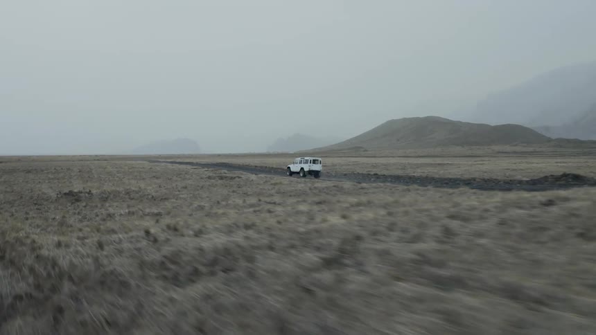 An aerial view of a white vehicle driving on a dirt road on an overcast day