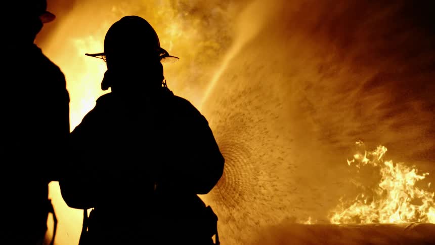 Two firefighters are holding a fire hose and spraying water towards flames