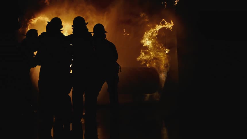 Firefighters standing together in the darkness holding a hose as it shoots water towards a raging fire