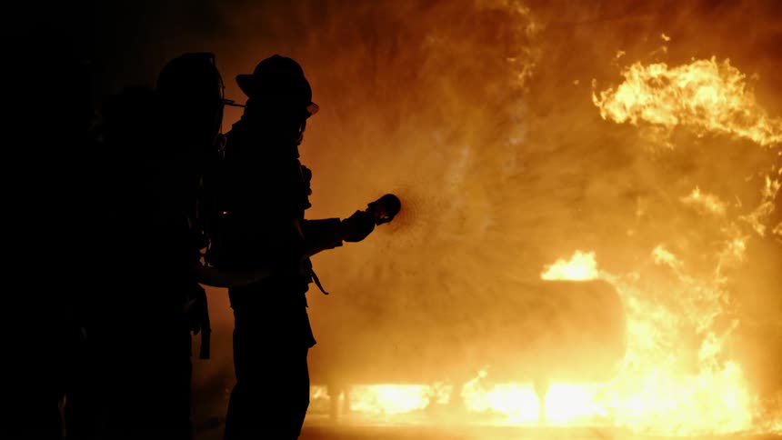 Two firefighters spray a hose towards a large burning fire