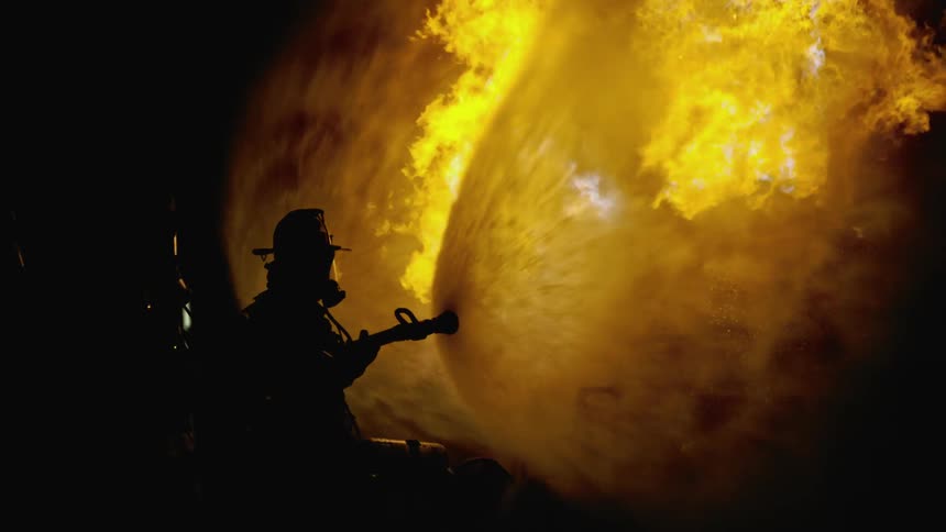 The silhouette of firefighter can be seen as they spray water at flames using a fire hose