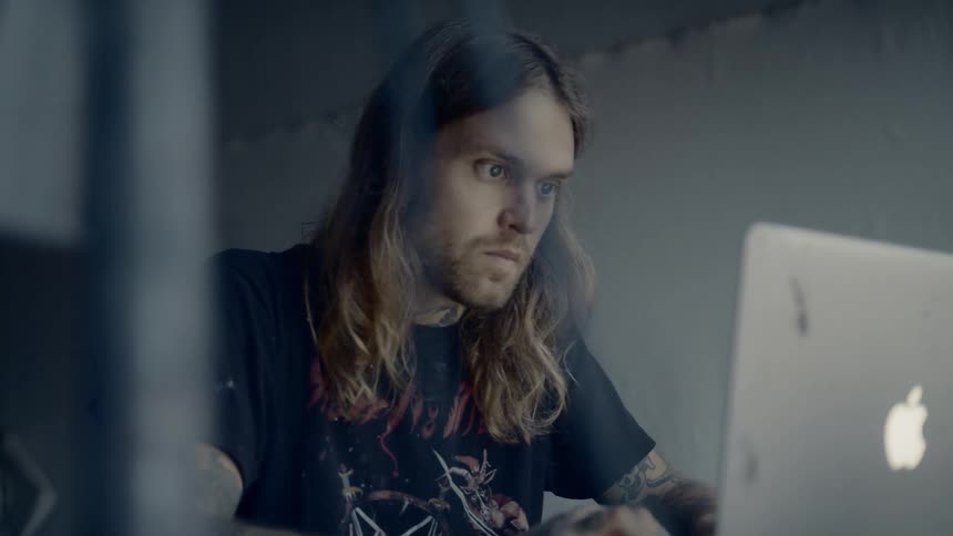 Long haired man sitting at the desk uses laptop in a room