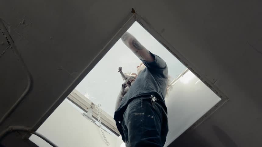 A young man with long hair holds a cat while he reaches up to open a skylight window