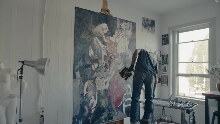 Man paints a large picture in a white room as he stands on a bench