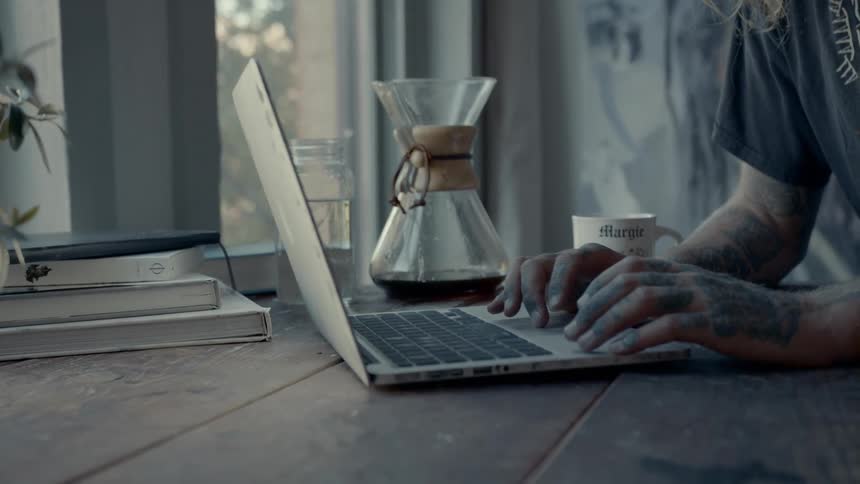 A tattooed man using a laptop attable next to a window