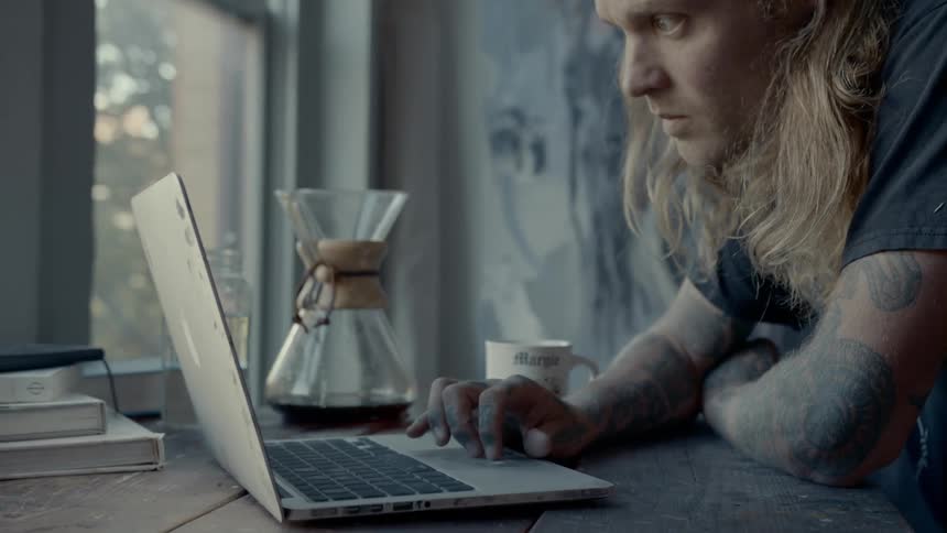 A man with tattoos scrolls on a laptop next to a carafe of coffee