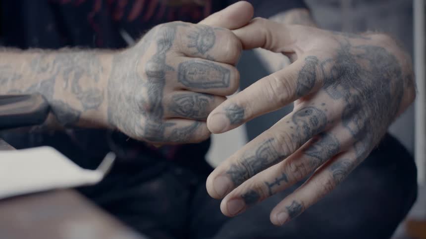A male with heavily tattooed hands and fingers sits hand while gripping his own thumb