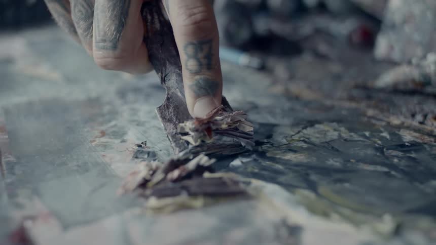 A man with tattoos on his fingers pushes a chisel across a flat surface to remove a layer of paint