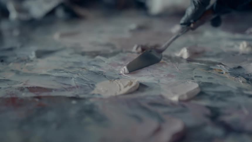 An artist wearing a black disposable glove carefully uses a metal tool to spread blobs of paint evenly onto a smooth surface