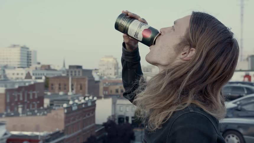 A man standing on a roof top parking lot tilts his head back as he drinks a beer