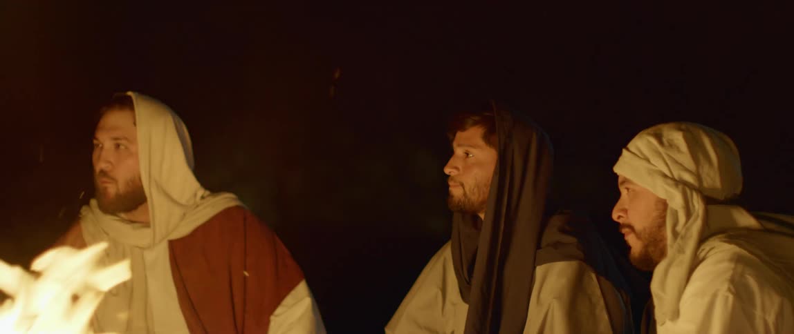 A group of men with robes sit beside a fire outside