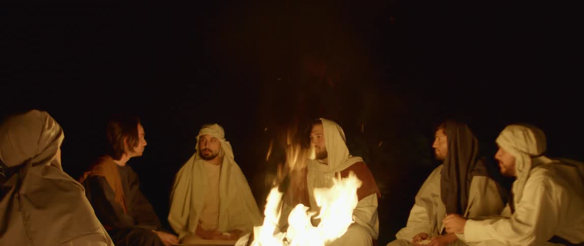 A group of men wearing costume robes sit around a fire