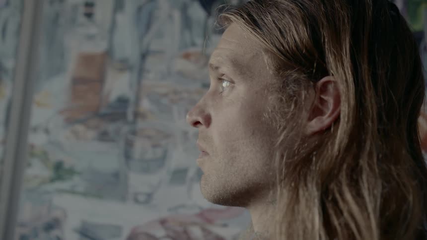 Long haired blonde man standing next to the painted wall looks ahead and drops his gaze
