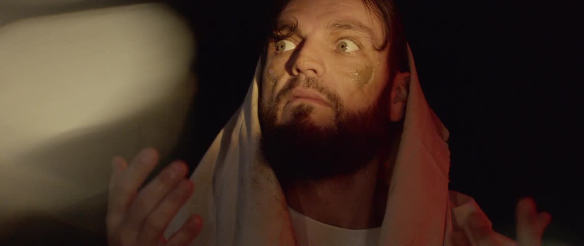 A man with dirt on his face and a sheet over his head looks at his hands and looks around