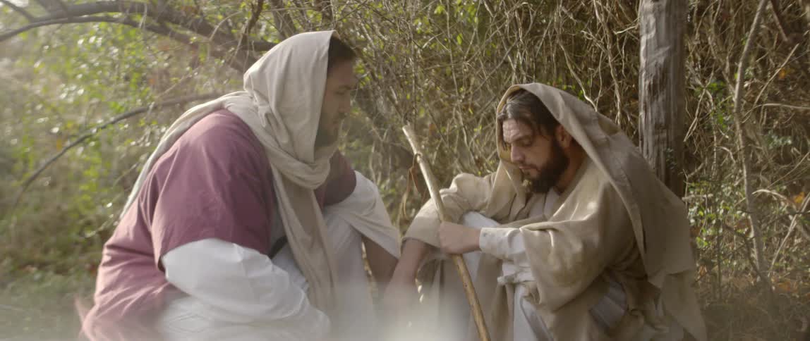 A man in a cloak sits on the ground and another man comes up and bend down to talk with him