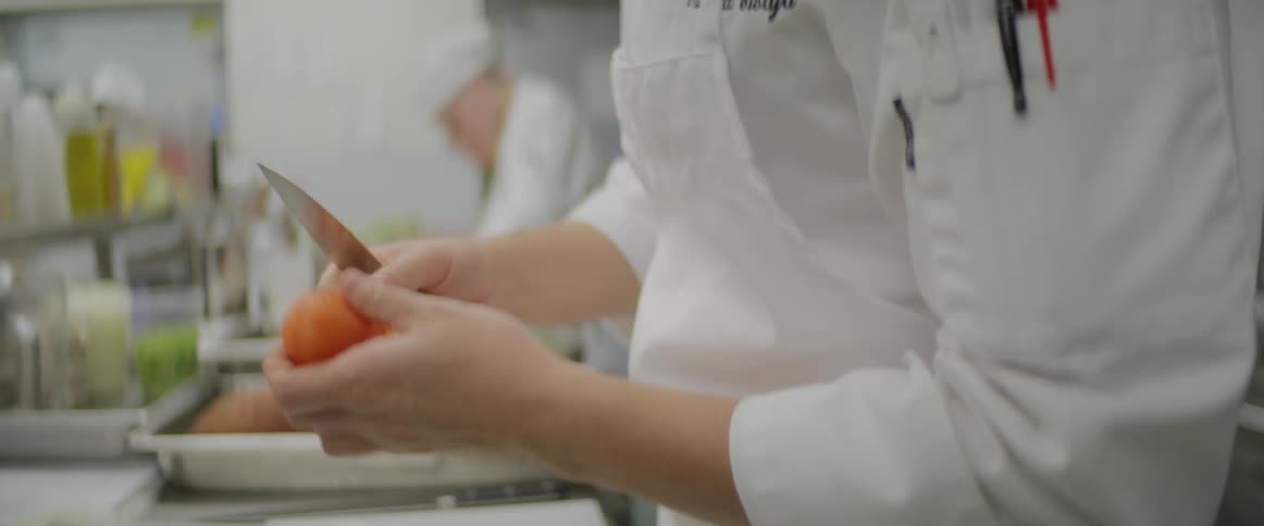 In a prep kitchen a chef carefully peels a small tomato with a knife