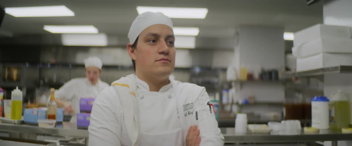 A chef places a hand towel over his should and stands with his arms folded across his chest within a large prep kitchen